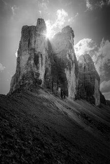 Le Tre Cime di Lavaredo fotografate in bianco e nero dal basso, con il sole che filtra tra le pareti rocciose e ne evidenzia la verticalità e la maestosità. Il versante in primo piano è scuro e pietroso, mentre il cielo luminoso incornicia le forme austere della montagna.