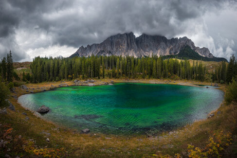 Il Lago di Carezza nelle Dolomiti riflette le acque turchesi mentre le cime rimangono in parte nascoste dalla nebbia. Il cielo nuvoloso e la luce soffusa creano un